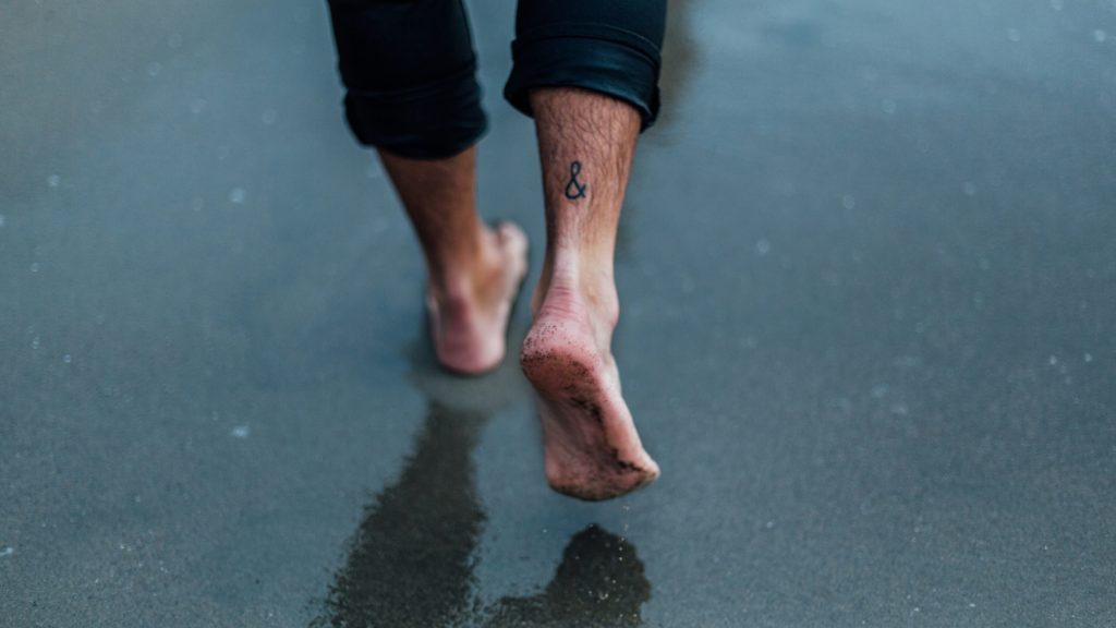 A person walking barefoot. Image courtesy of Unsplash.com. https://unsplash.com/photos/person-with-tattoo-on-foot-walking-on-wet-sands-orFHTN5BBtM