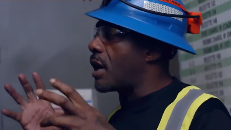 Idris Elba wearing a blue safety hat in a gold mine.