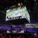 League of Legends World Championship game play being shown on the over head screens of the venue with players underneath and crowd cheering