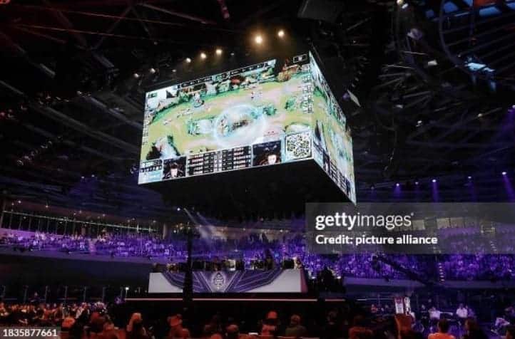 League of Legends World Championship game play being shown on the over head screens of the venue with players underneath and crowd cheering