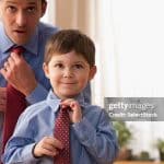 A dad teaching his son to tie a tie. It is an example of a parenting style.