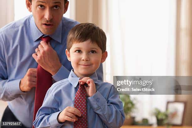 A dad teaching his son to tie a tie. It is an example of a parenting style.