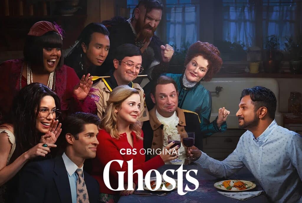 Screenshotted promotional image of the paramount plus tv show Ghosts with the two main cast members at a dinner table surrounded by the ghosts on one side