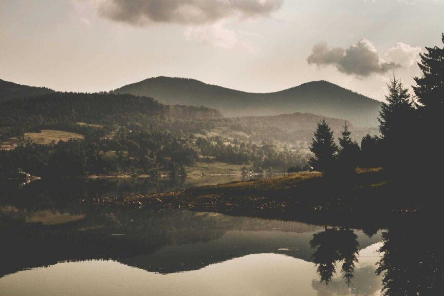 Fog palette tones define this serene mountain scene—misty peaks, soft lake reflections, and a cloudy sky create a calm, dreamlike mood. Image by Vlad Bagacian in Courtesy of Pexel