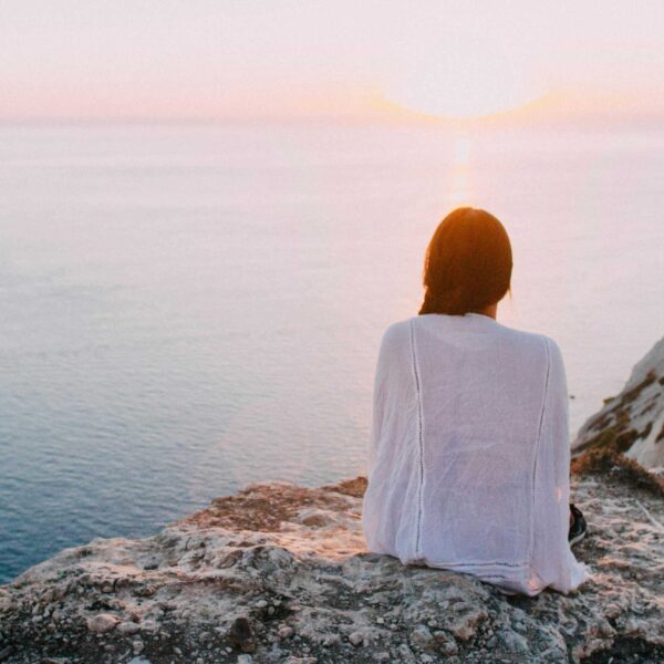 A serene view of a woman sitting on a rocky cliff, gazing at the ocean during sunset. Perfect for relaxation and travel themes. Mental Health