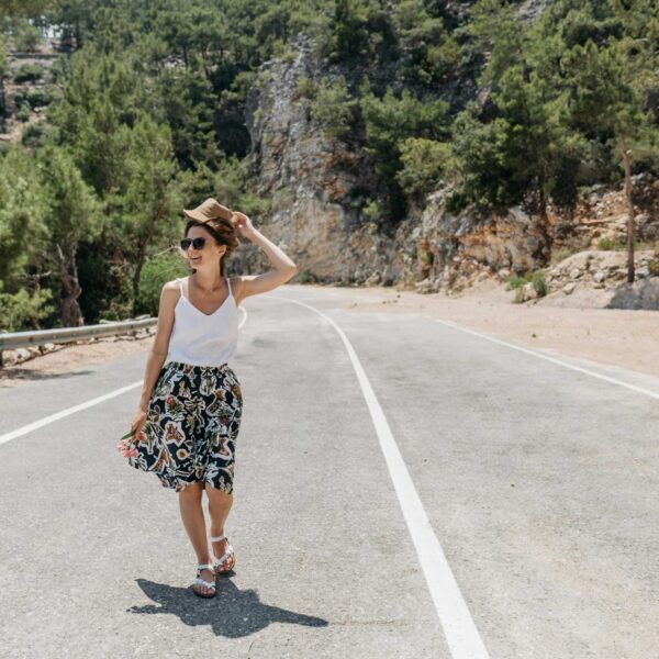 A woman smiling and walking on a sunny road surrounded by trees and nature with summer accessories
