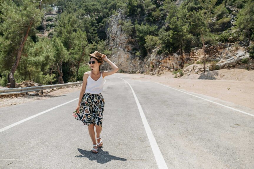 A woman smiling and walking on a sunny road surrounded by trees and nature with summer accessories