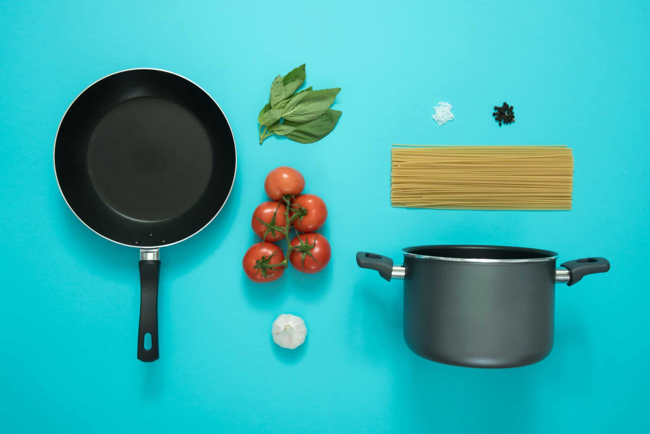 Flat lay photo featuring a frying pan, pot, tomatoes, and spaghetti on a blue background.