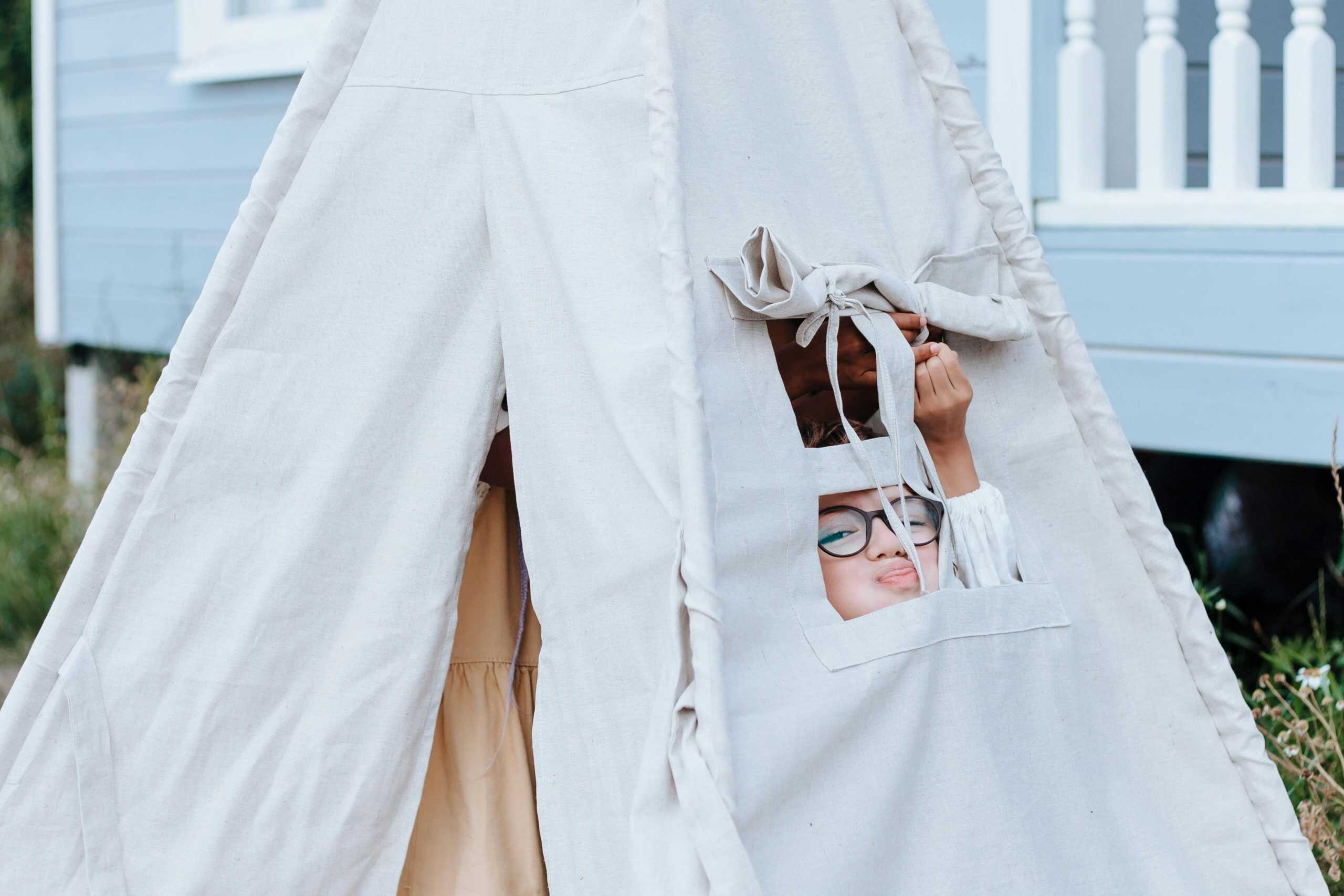 A young child with glasses peeks out from a teepee tent set outdoors, embodying the joy of play and exploration. Backyard, cheap family activities