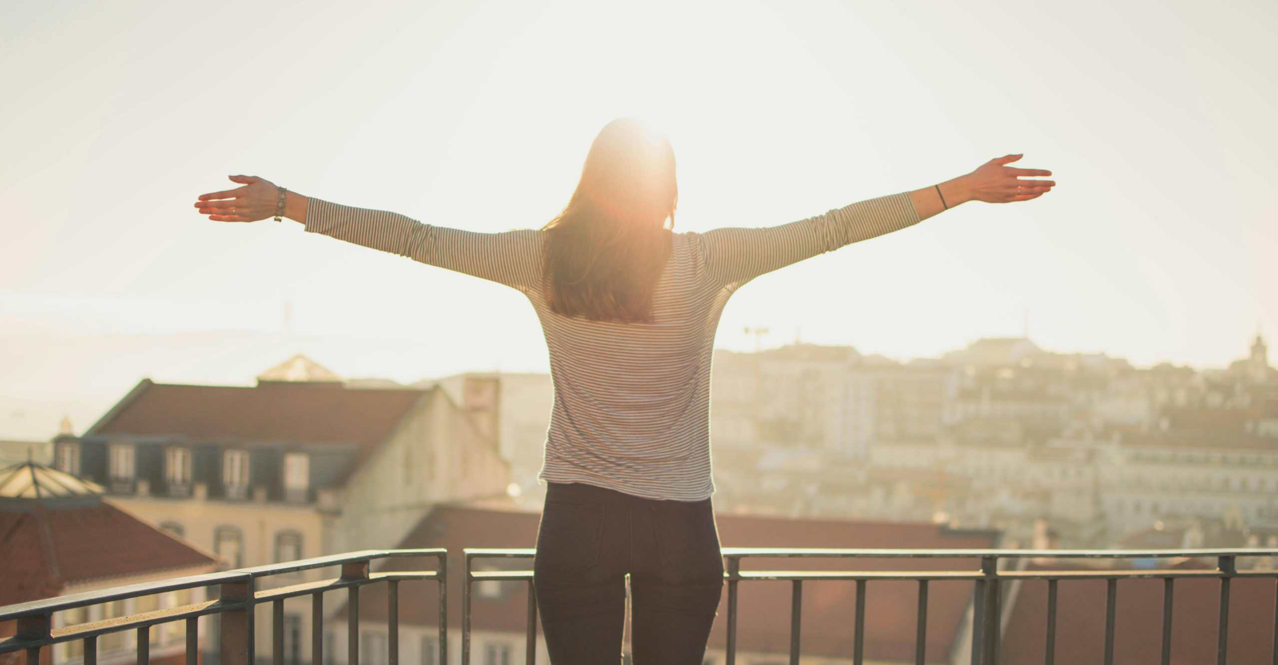 A woman stands with outstretched arms on a sunny balcony, embracing the morning light. Morning routine