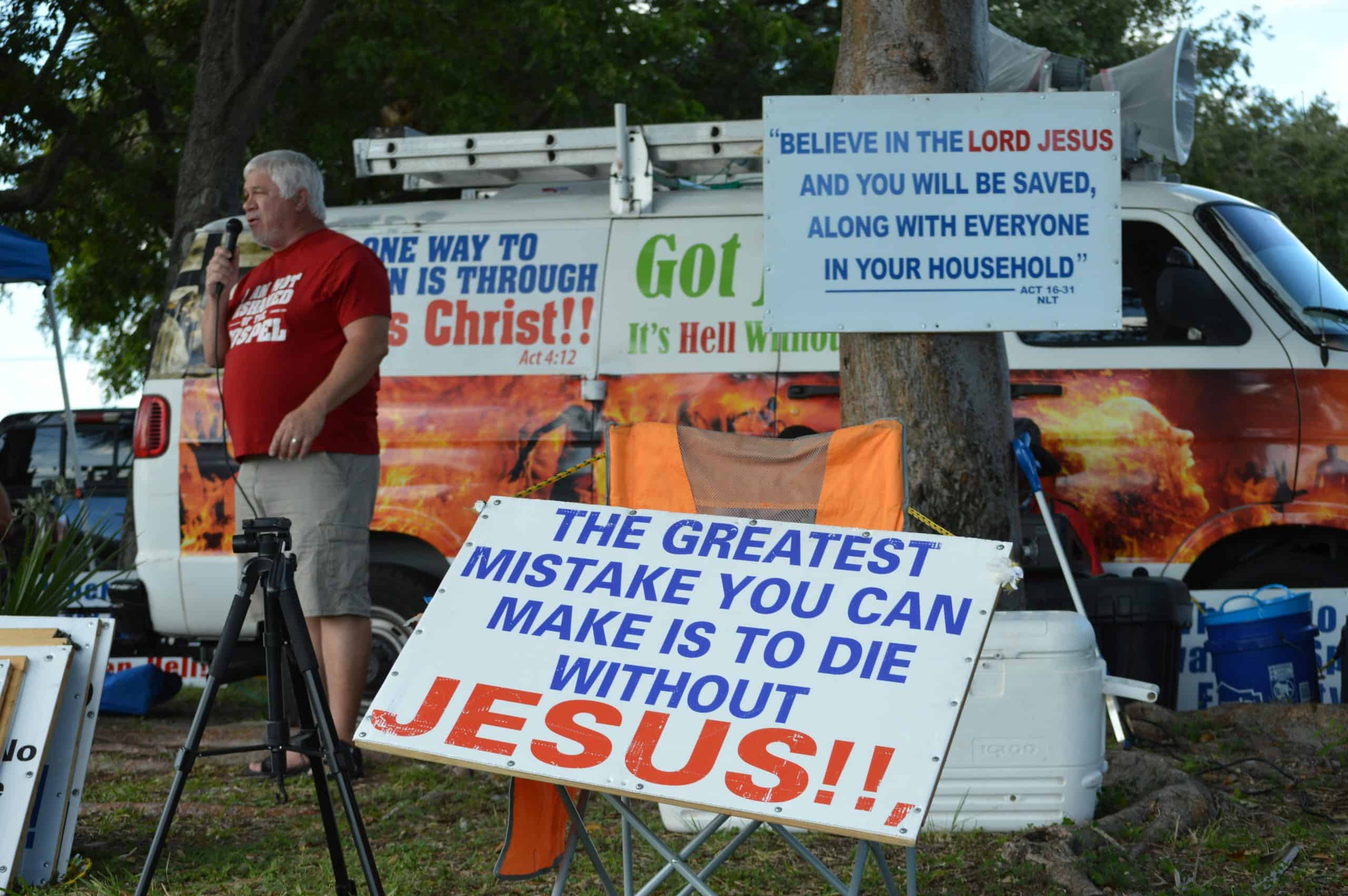 A man engages in outdoor evangelism with religious signs and a microphone by a van, daily devotion