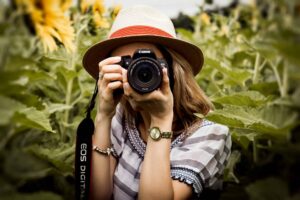 Woman with hat practicing travel photography in a summer sunflower field using a Canon camera. Image credit: by Andre Furtado via Pexels