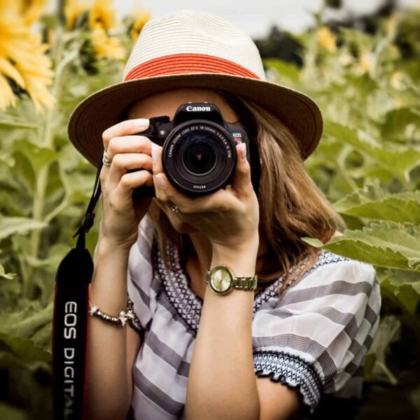 Woman with hat practicing travel photography in a summer sunflower field using a Canon camera. Image credit: by Andre Furtado via Pexels