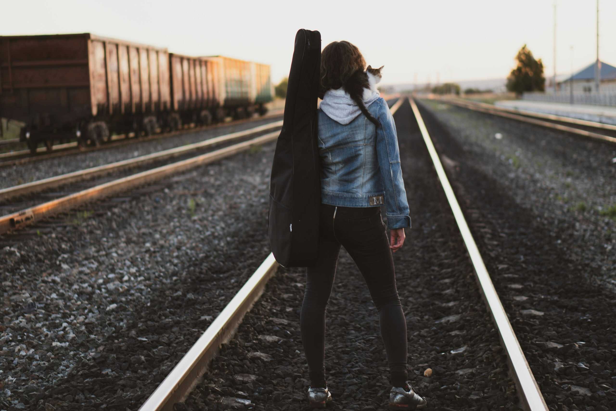 Woman with guitar case stands on railway tracks, looking towards the horizon at sunrise.