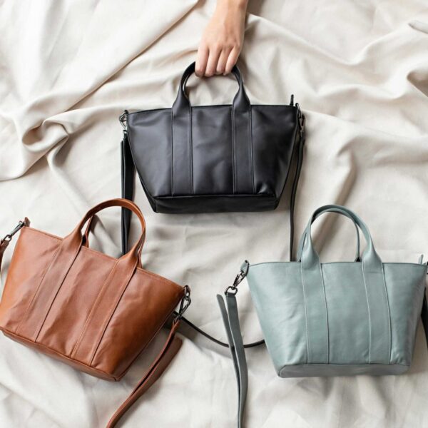 Top view of three stylish leather designer bags in black, brown, and gray on a soft fabric surface.