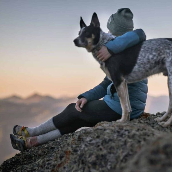 A person wearing a beanie and winter jacket sits on a rocky mountain with a dog, overlooking sunset views. pets