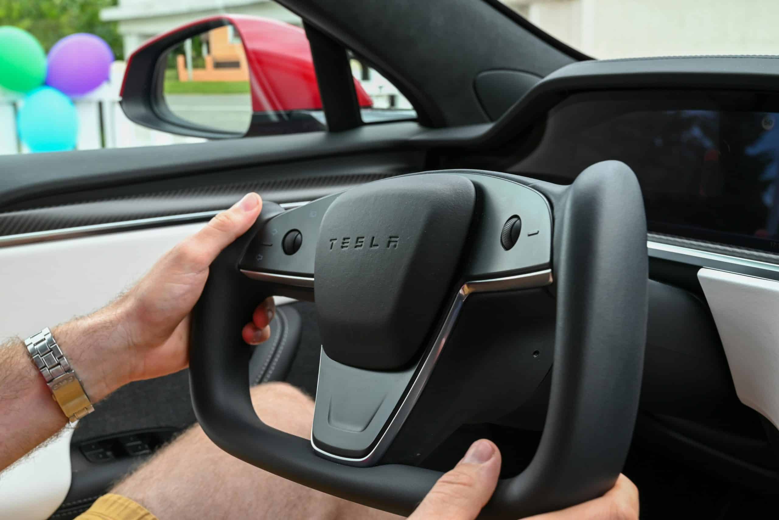 Hands gripping Tesla's minimalist yoke steering wheel in an electric vehicle interior.