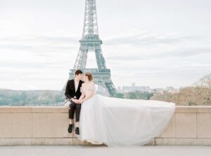 This photograph features a couple dressed for a wedding. They are sitting in front of the Eiffel Tower in Paris. Paris can be included in a list of honeymoon destinations.