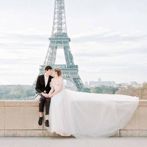This photograph features a couple dressed for a wedding. They are sitting in front of the Eiffel Tower in Paris. Paris can be included in a list of honeymoon destinations.