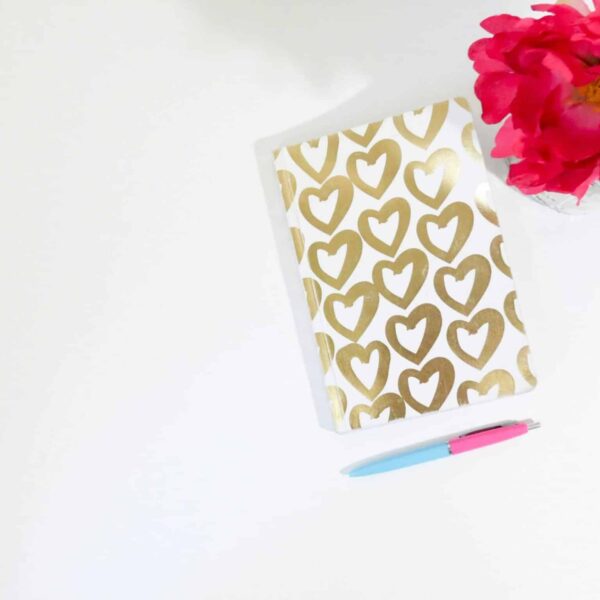 This photo features a journal with gold drawn hearts on the cover. There photo also contains flowers and a pen. Journaling can be a great way to develop a positive connection with yourself.