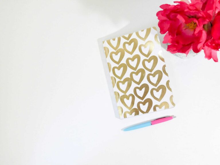 This photo features a journal with gold drawn hearts on the cover. There photo also contains flowers and a pen. Journaling can be a great way to develop a positive connection with yourself.