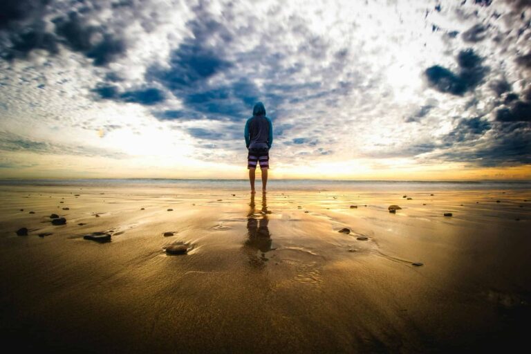 Cause to Pause: A lone figure stands on a tranquil beach during a vivid sunset, reflecting on the wet sand.