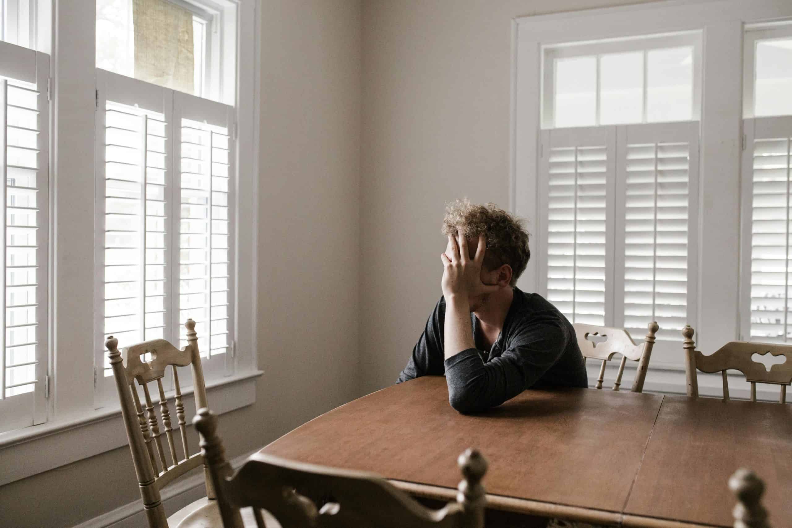 A man sits alone at a table in a bright room, displaying deep contemplation. mental health