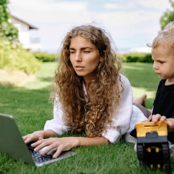 This photograph features a woman on a laptop. A kid is next to her with a toy. Parenting can include a balance of screen time and alternative activities.