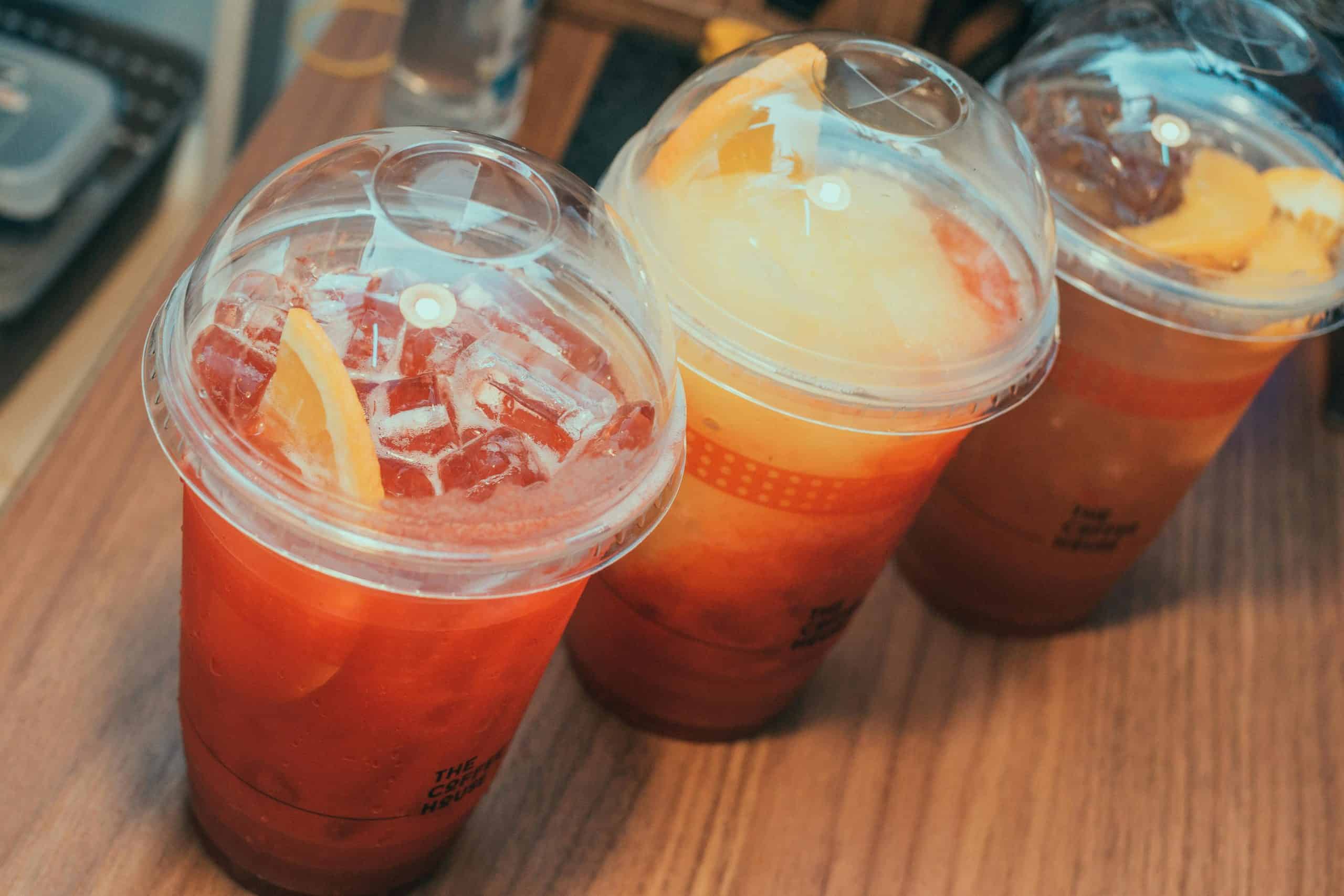 Close-up of three iced fruit drinks in plastic cups with sliced fruit garnish on a wooden surface.