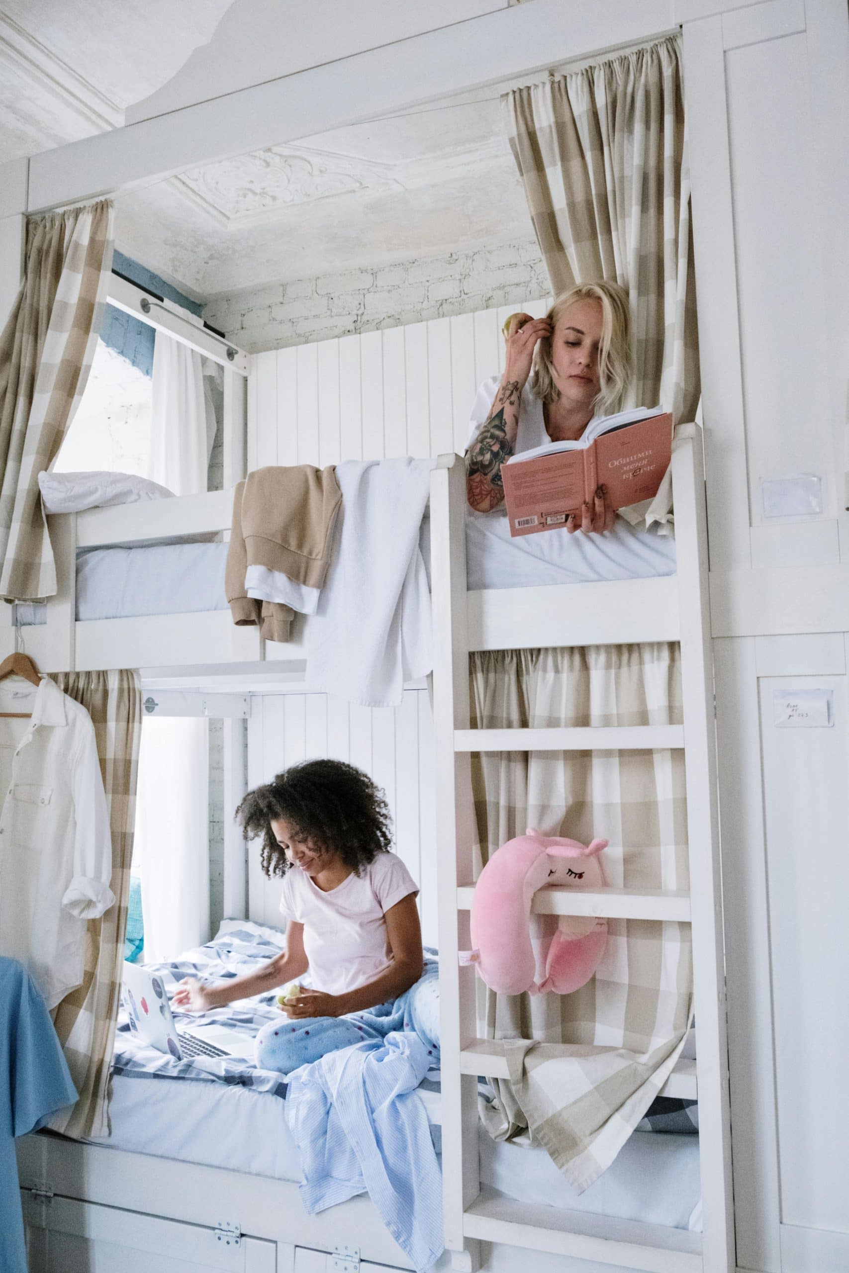 Two women enjoying a cozy bunk bed setup, reading and using a laptop, in a bright room with neutral tones.