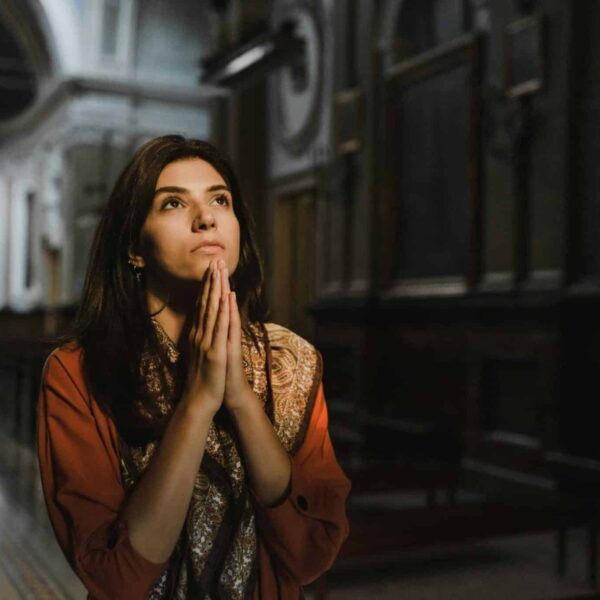 A woman is praying with hands clasped in a dimly lit church, creating a contemplative mood. Daily devotional