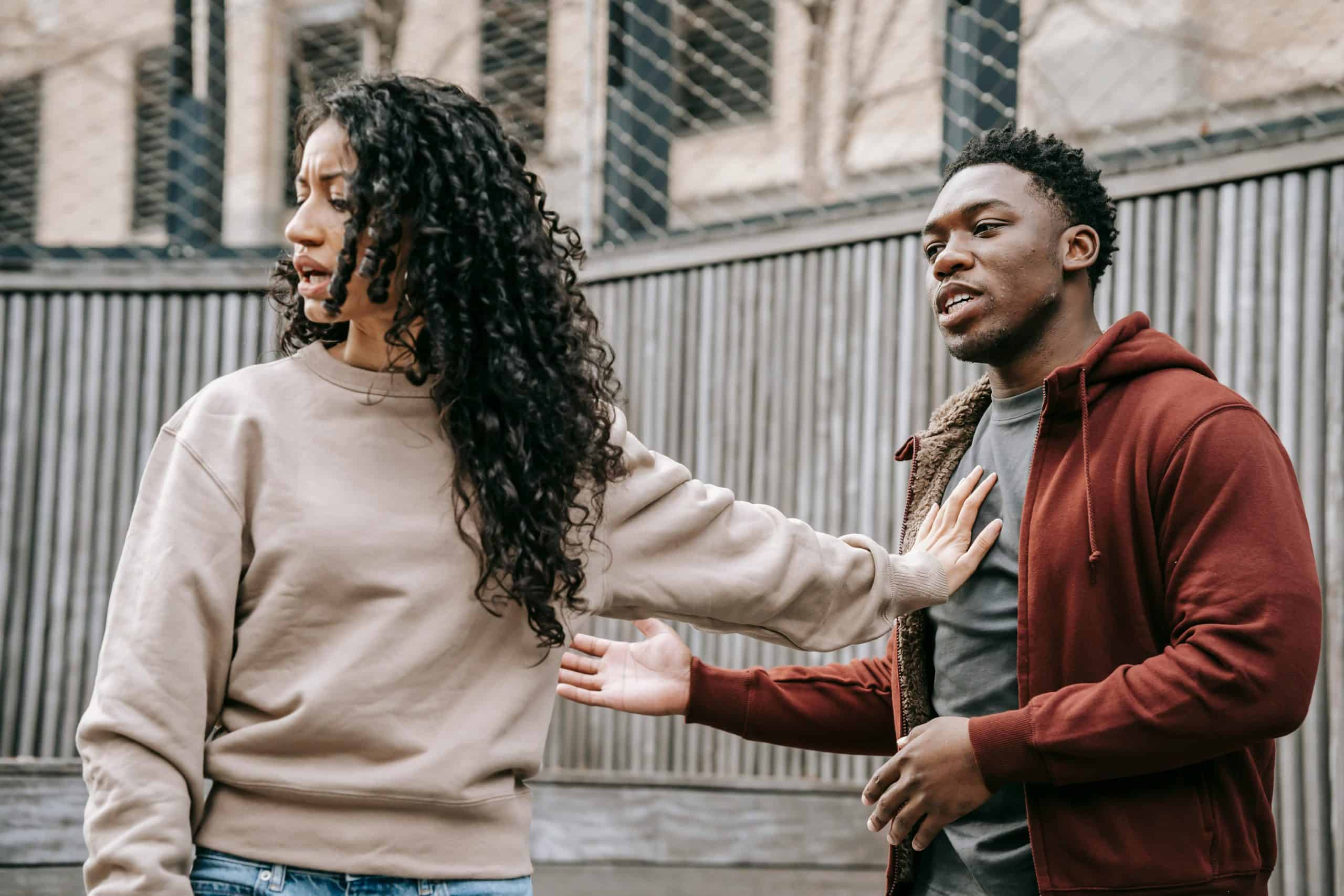 A couple in casual clothing appears to be in a disagreement outdoors with grey fencing background. sabotaging, relationship