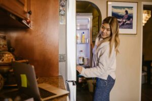 A woman at home checks her refrigerator while working on a laptop in a cozy setting. leftovers