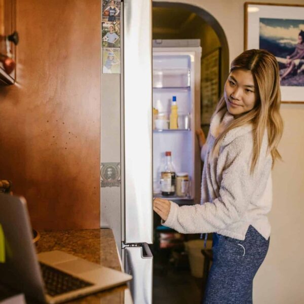 A woman at home checks her refrigerator while working on a laptop in a cozy setting. leftovers