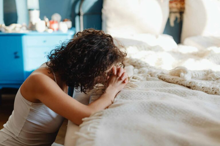 A woman kneels in contemplation by her bed, showcasing spirituality and tranquility in a warmly lit bedroom. morning routine