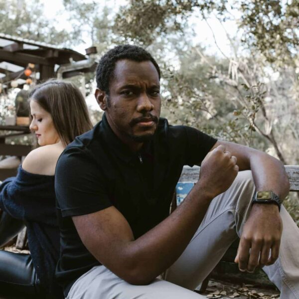 A thoughtful man sits outdoors, with a woman sitting distantly behind him. emotionally unavailable