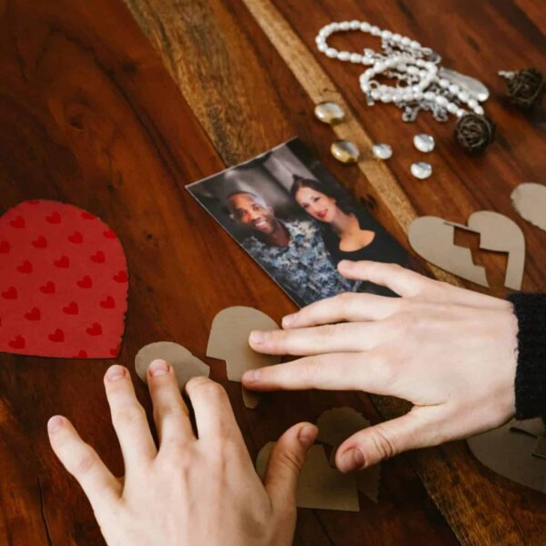 Close-up of hands arranging broken hearts near a couple's photo on a wooden table. Need healing after a breakup