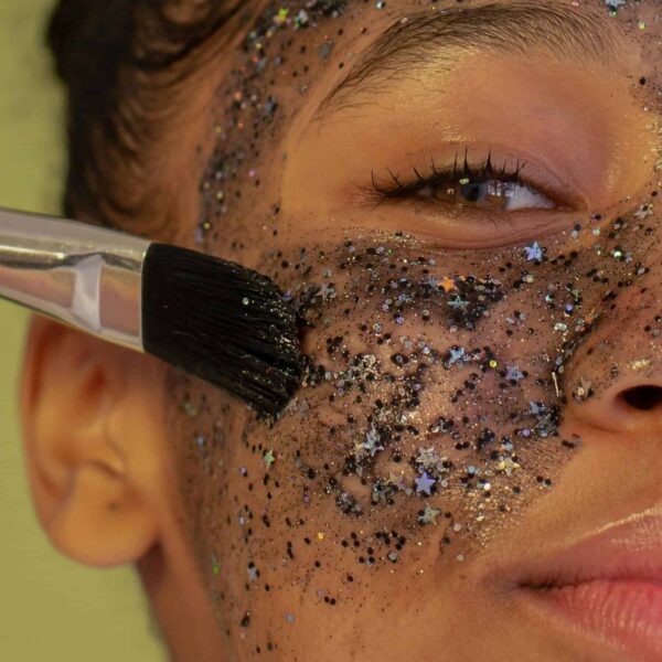 Crop unrecognizable young African American female with brush applying facial mask with glitters during beauty procedure and looking at camera on green background. Makeup hacks for Y2K