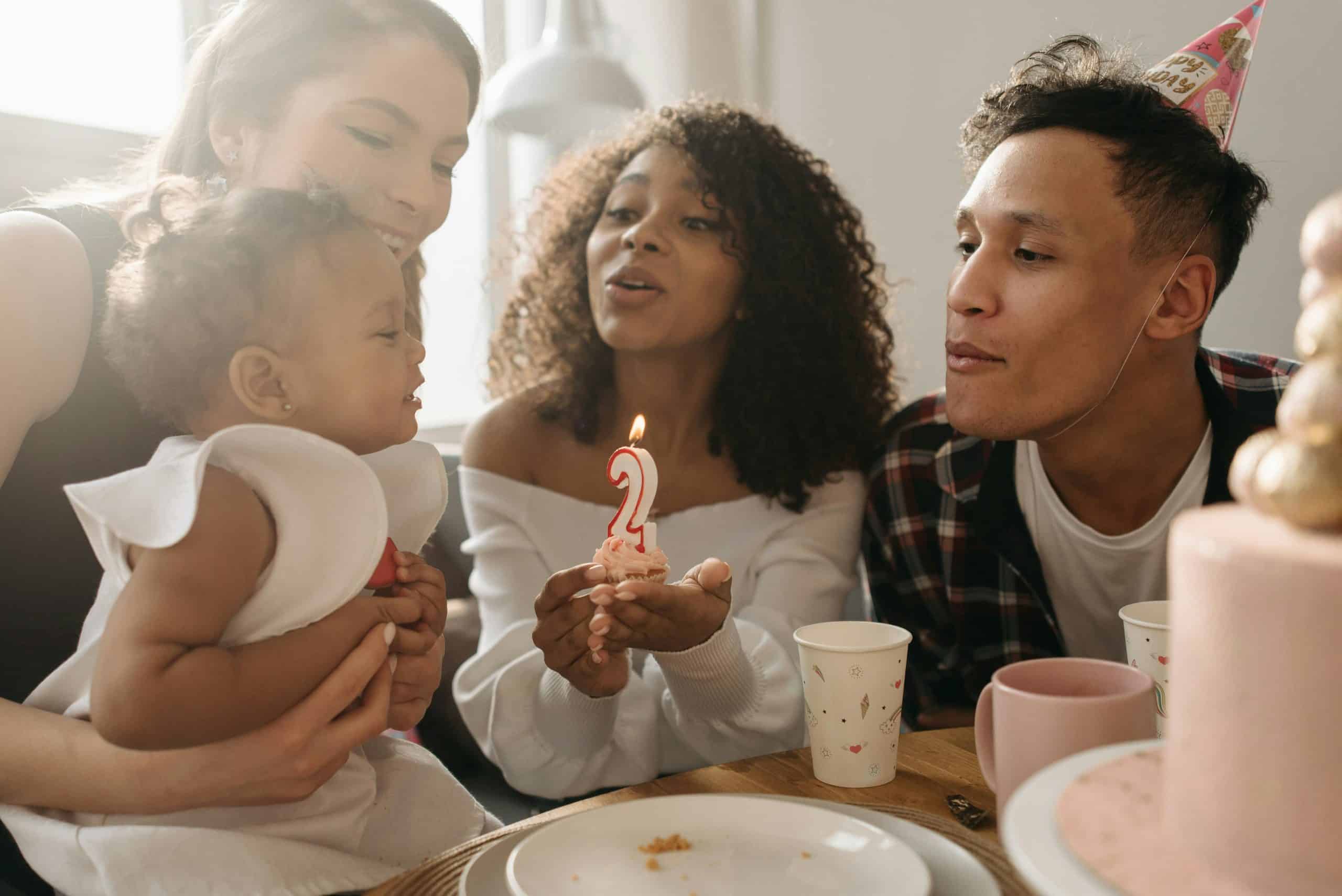 A diverse family celebrates a toddler's second birthday with a cupcake and candle indoors. Family traditions