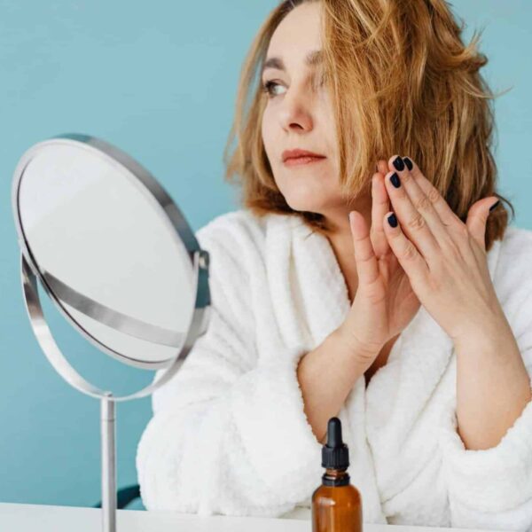 Woman in white robe applying skincare oil, looking in the mirror with manicured nails. beauty product launches