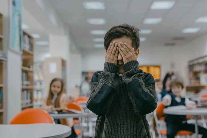 Young boy covers face in a bustling school classroom, indicating stress or anxiety.