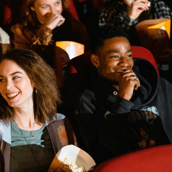 Group of diverse friends laughing and enjoying popcorn while watching a movie in a theater.