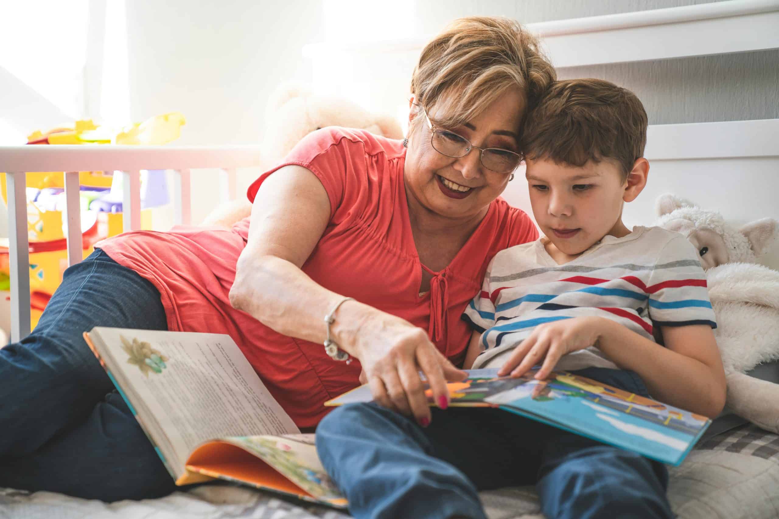 Elderly woman reading a book with her grandson on a cozy bed in a sunlit room.