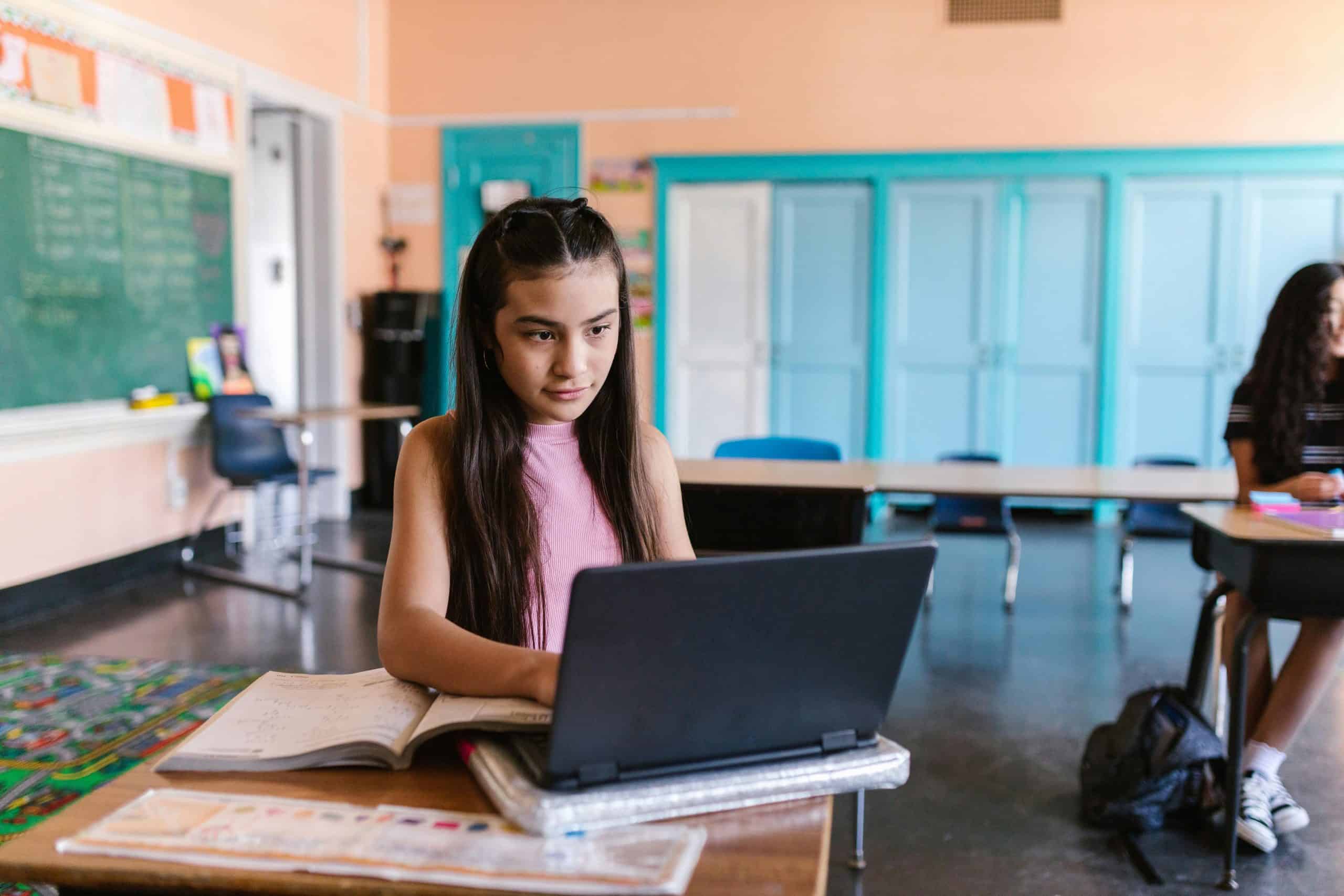 A focused young girl using a laptop for schoolwork in a bright classroom environment. back to school tech