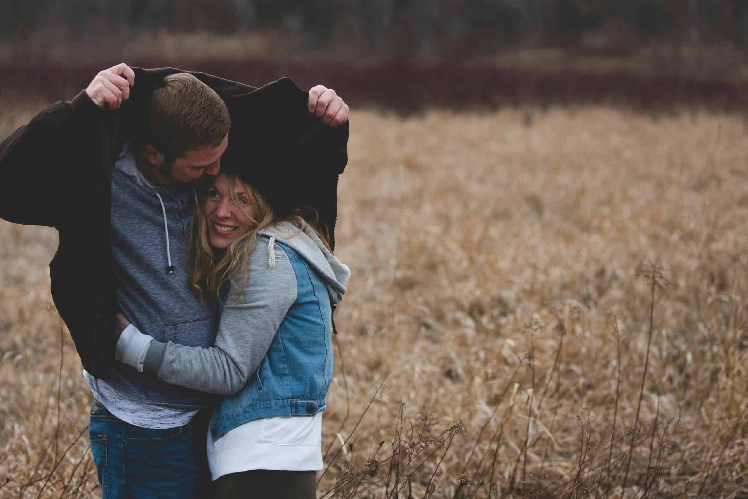 A couple shares a warm embrace under a coat in a rustic autumn field, expressing love and togetherness. interdependence