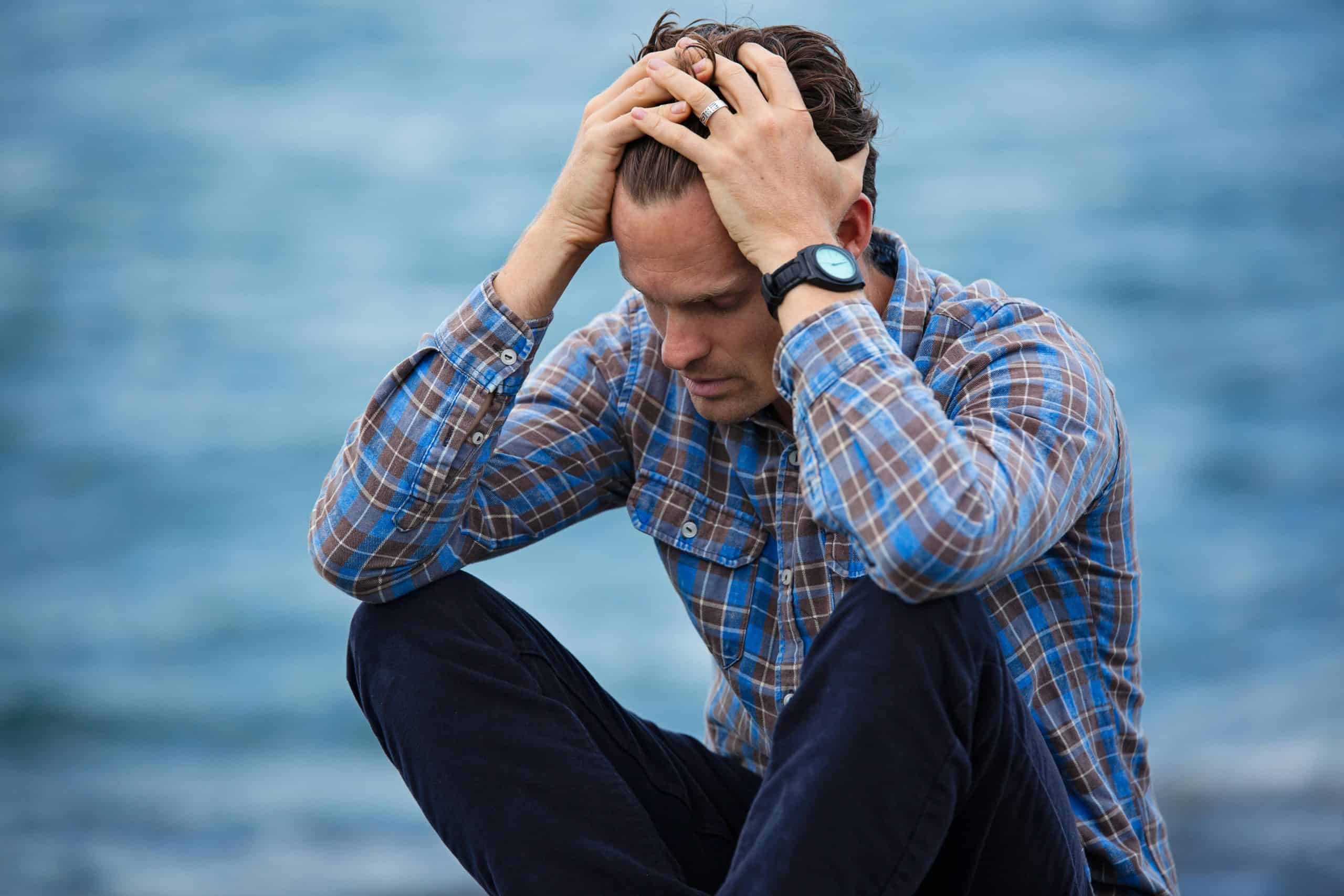 Trust God's Timing. A man in a plaid shirt sits by the water looking distressed, symbolizing stress.