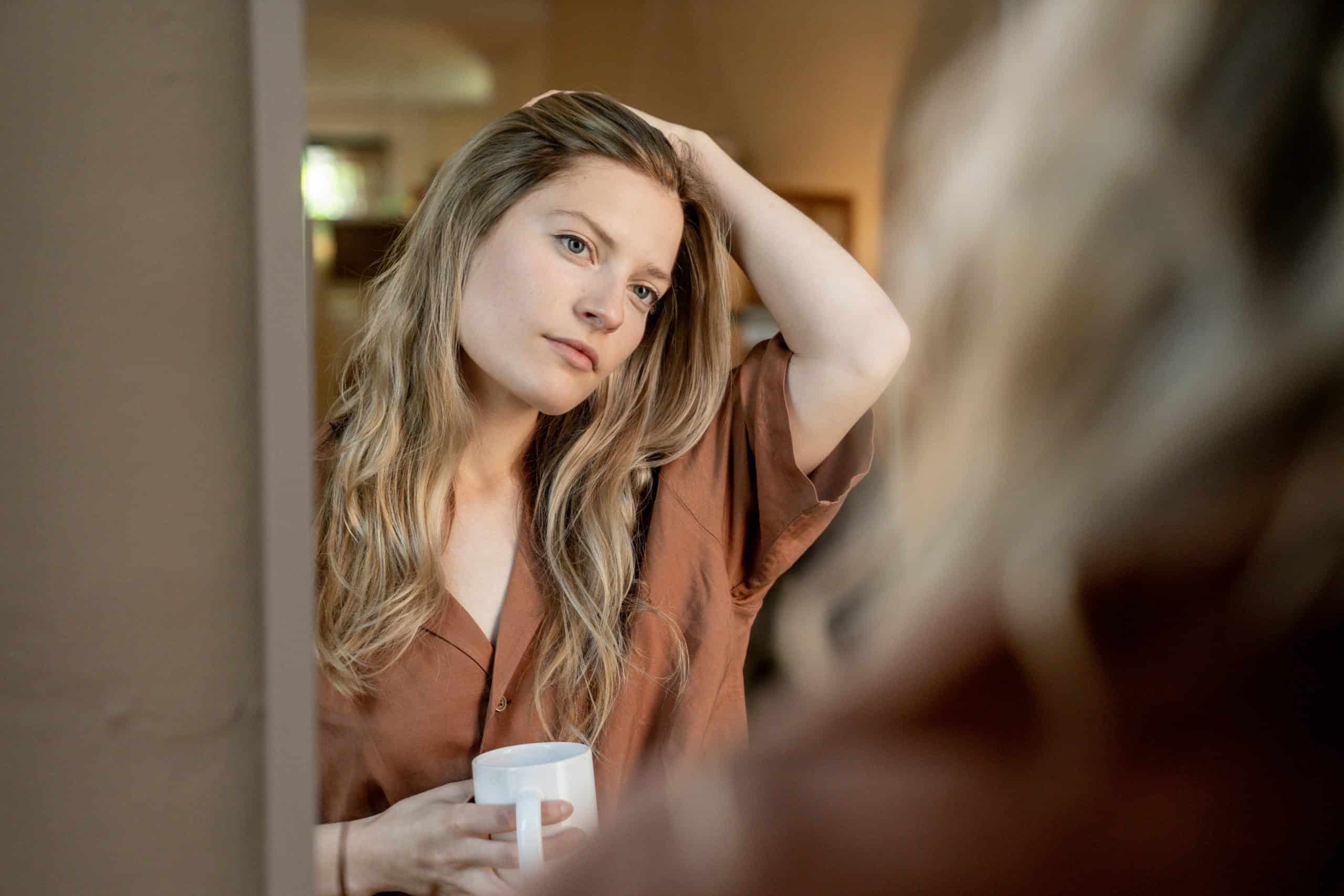 This photo is of a woman looking into a mirror and holding a coffee mug. You can check out your reflection throughout your day and use oil-blotting sheets as needed.