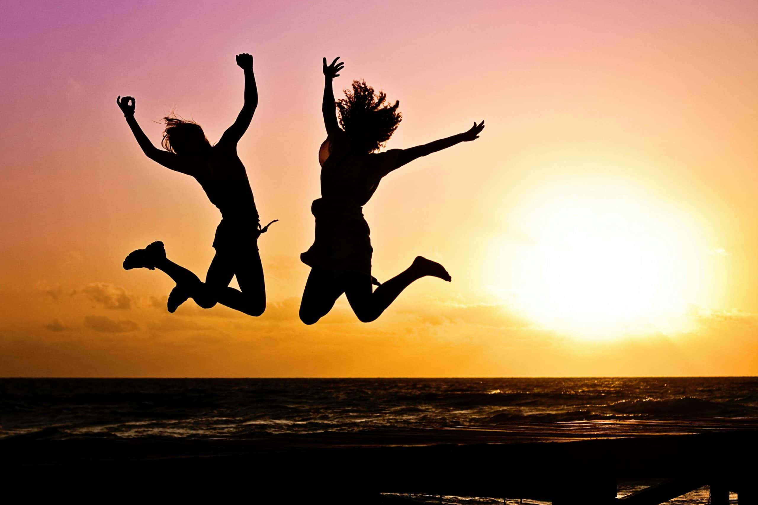 Silhouettes of two people joyfully jumping on a beach during a vibrant sunset. daily devotion