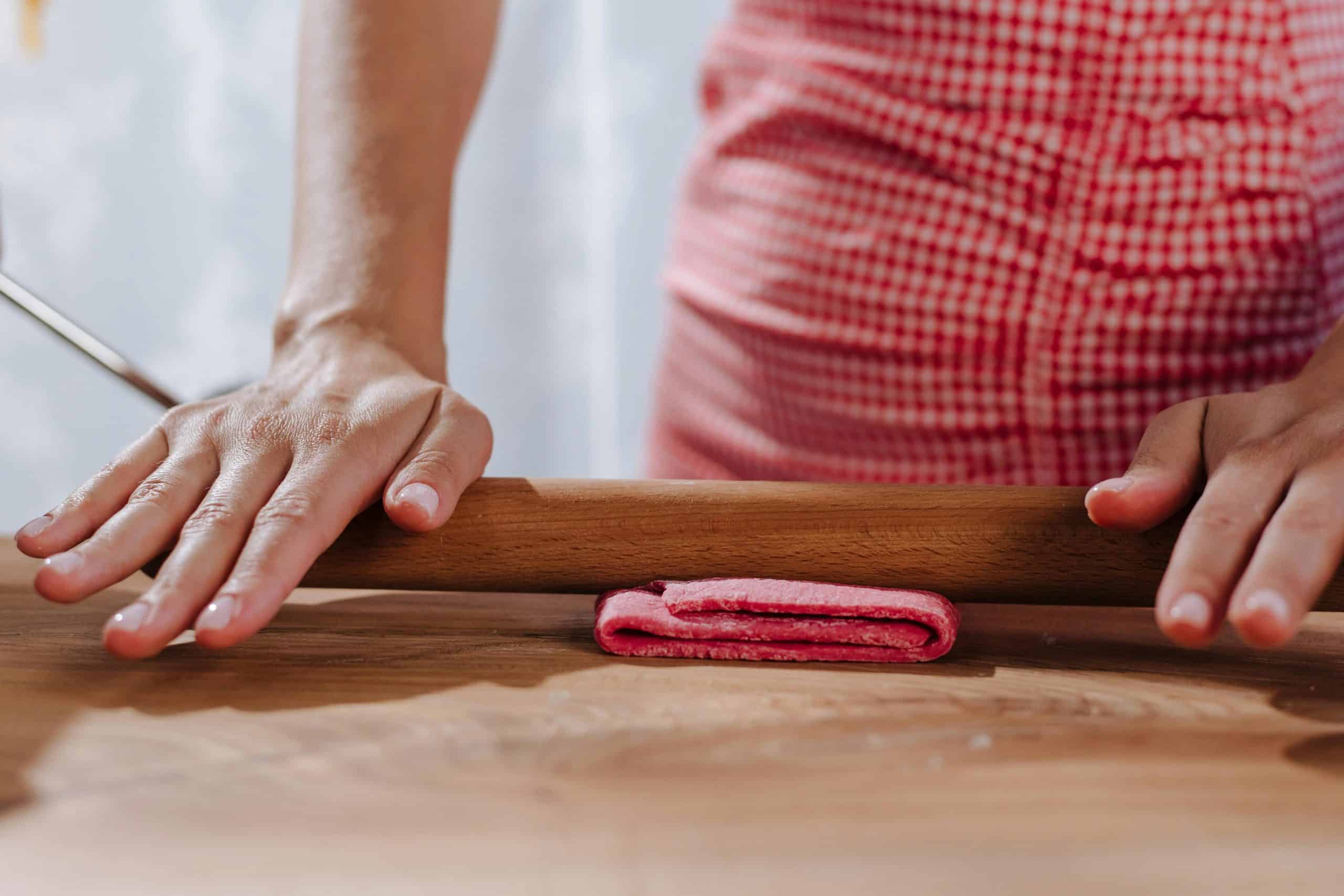 Close-up of hands using a rolling pin to prepare pasta on a wooden surface in a kitchen.