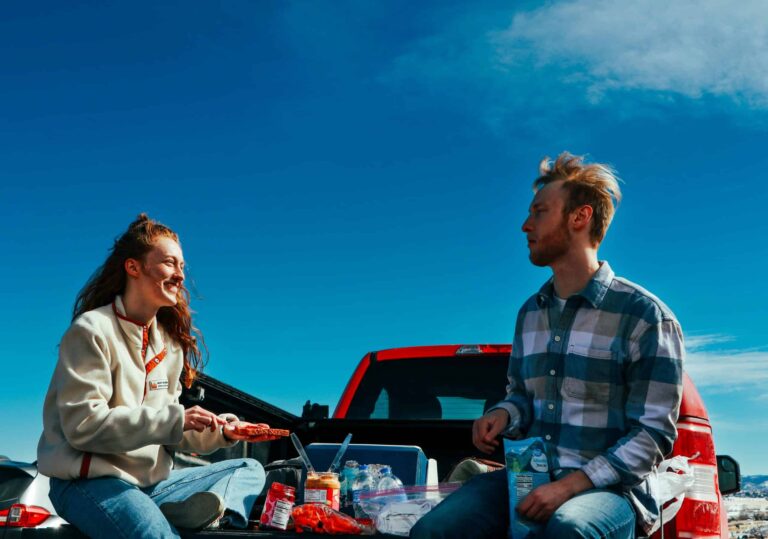 A cheerful couple having a picnic on a truck tailgate under clear blue skies. game day